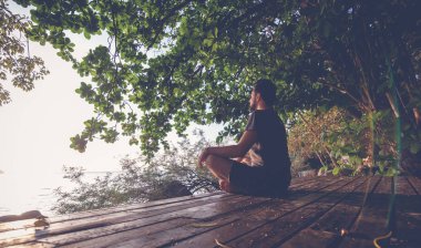 Genç adam gün batımında, deniz manzaralı niteliğine ahşap bir platform üzerinde meditasyon retro tonlarda vurdu