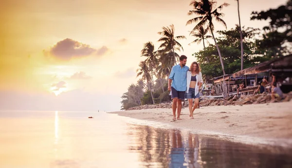 若く美しい愛の新婚旅行の休暇旅行中に夕暮れビーチで腕を組んで歩いて幸せなカップル — ストック写真