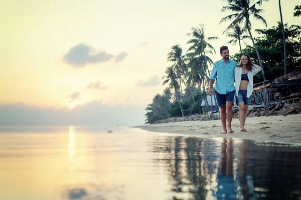 若く美しい愛の新婚旅行の休暇旅行中に夕暮れビーチで腕を組んで歩いて幸せなカップル — ストック写真