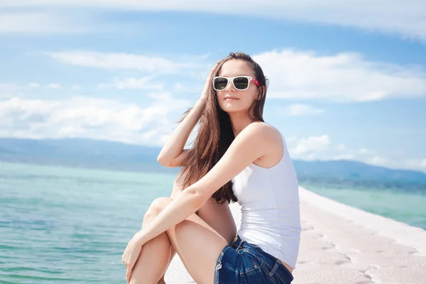 サングラス白の シャツとデニムのショート パンツ ブルーの熱帯の海の海岸にでスタイリッシュな美しい女性ブルネットの少女 — ストック写真