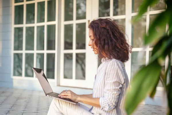 Молодая красивая женщина сидит на террасе с ноутбуком в фоновом режиме дома . — стоковое фото