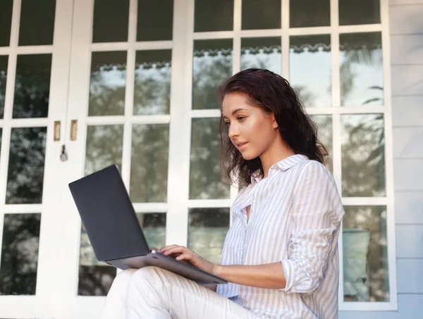 Молодая красивая женщина сидит на террасе с ноутбуком в фоновом режиме дома . — стоковое фото