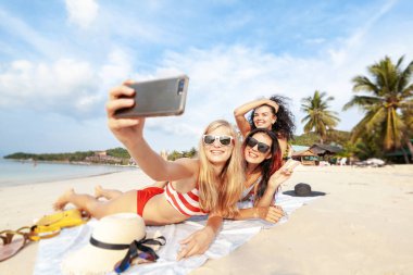 Beautiful happy girlfriends having fun on the beach taking selfies during vacation and travel clipart