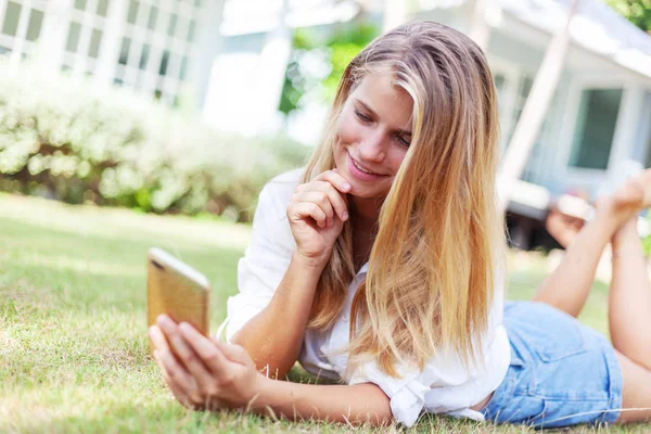 Menina loira bonita, em um gramado verde de verão, com um smartphone em suas mãos goza do sol — Fotografia de Stock