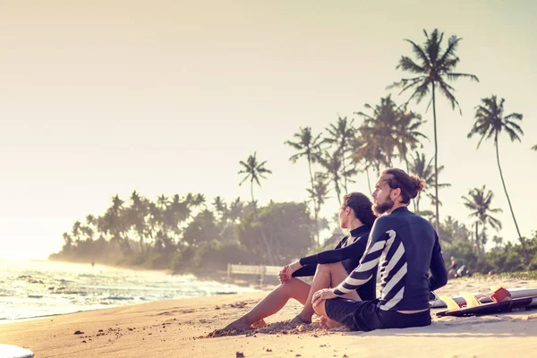 Young beautiful loving couple friends are sitting on the sand by the ocean with surfboards — 스톡 사진