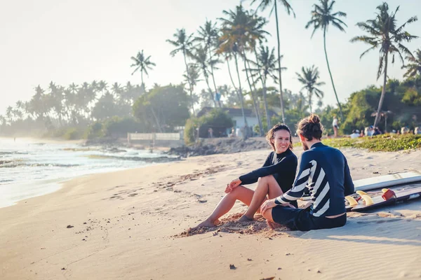 Jovens e belos amigos amorosos estão sentados na areia junto ao oceano com pranchas de surf — Fotografia de Stock