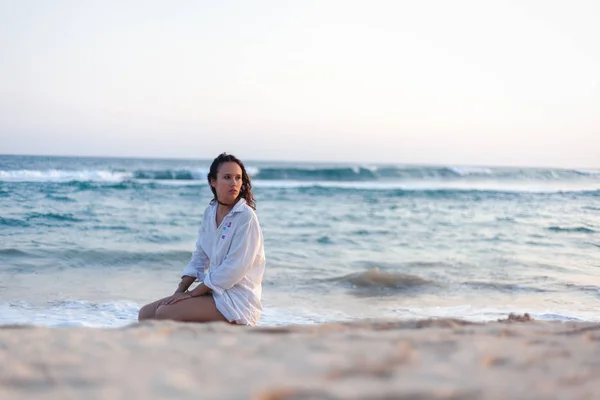 Hermosa joven morena con una camisa blanca sentada en el océano al atardecer —  Fotos de Stock