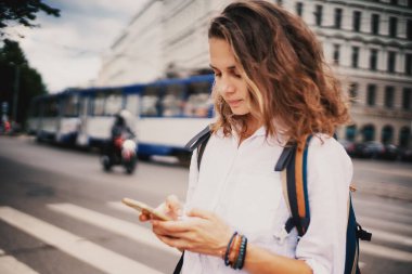 Beyaz gömlekli genç bir kadın, yaya geçidine yakın bir yerde ulaşımı olan bir caddedeki akıllı telefona, navigasyon başvurularına, haritalara, toplu taşıma programlarına bakıyor.