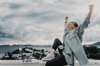 Güzel, heyecanlı, şoke olmuş, şehir manzaralı, gözlüklü, dizüstü bilgisayar kullanan genç ve mutlu bir iş kadınının görüntüsü..