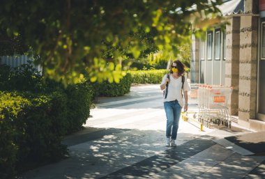 Koruyucu maskeli ve güneş gözlüklü genç bir kadın şehirde bir yaz caddesi boyunca yürüyor, yeni normal bir yaşam tarzı.
