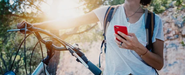 Una mujer está de pie con una bicicleta y un teléfono inteligente en sus manos en el bosque, mirando la navegación. Paseos en bicicleta — Foto de Stock