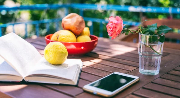 Wooden table on the sunny summer terrace. Open book, fruit plate and smartphone — Stock Photo, Image