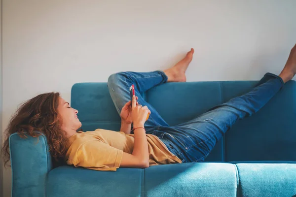 Jovem Bela Mulher Feliz Encaracolado Deitado Sofá Azul Usando Smartphone — Fotografia de Stock