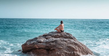Bikinili genç bir kadın kumsalda büyük bir taşın üzerinde oturur ve denize bakar, doğayla rahatlar ve meditasyon yapar..