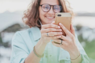 Gözlüklü güzel, mutlu, kıvırcık kız şehrin manzaralı akıllı telefon ekranına bakıyor. Modern teknolojiler ve şehir yaşam tarzı.
