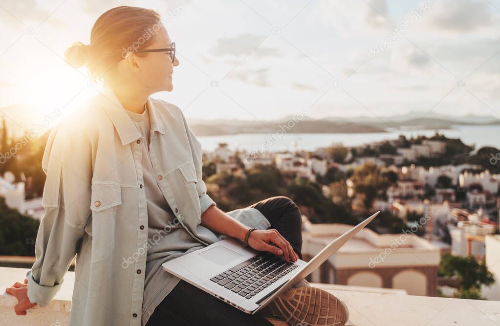Beautiful curly young girl with glasses working on a laptop with a city view at sunset. Modern technologies and urban lifestyle. Freelance and online education