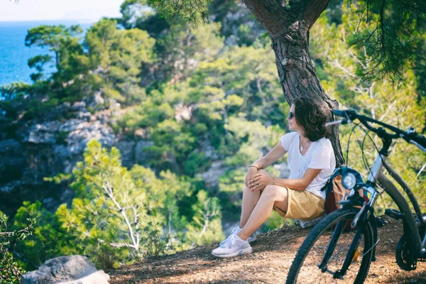 The girl has a rest in the forest with a bicycle, enjoys nature and loneliness. Summer vacation and travel by bicycle, sports and outdoor activities.