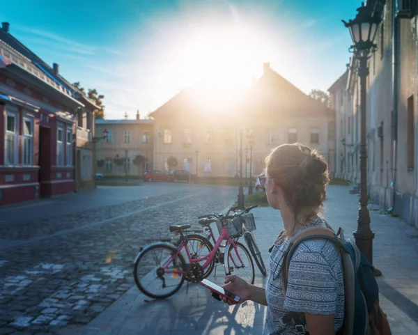 ヨーロッパの街の路上でスマートフォンを手にした若い女性 ヴィンテージの建物や駐車自転車 都市のコンセプトでの自転車レンタル — ストック写真