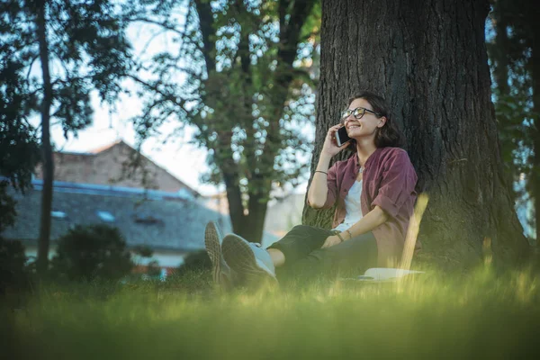 Młoda Kobieta Okularach Siedząca Parku Rozmawiająca Przez Telefon Komórkowy Opierająca — Zdjęcie stockowe