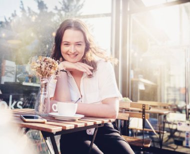 Genç, güzel, kıvırcık bir kadın yaz günü kafede kahve içiyor. Yüzünde mutlu bir gülümsemeyle.