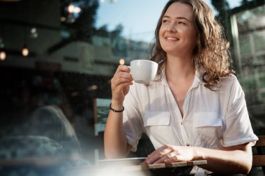 Genç, güzel, kıvırcık bir kadın yaz günü bir kafede kahve içiyor. Yüzünde mutlu bir gülümsemeyle..