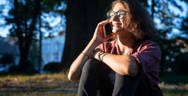 Parkta çimlerin üzerinde oturmuş cep telefonu kullanan güzel bir kız öğrenci..