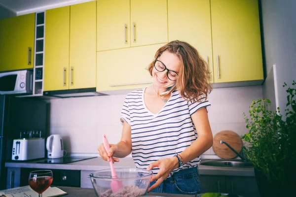 Güzel, mutlu, genç bir kadın mutfakta tatlı hazırlıyor. Cep telefonu, hobiler, eğlence ve yemek konseptini kullanıyor.