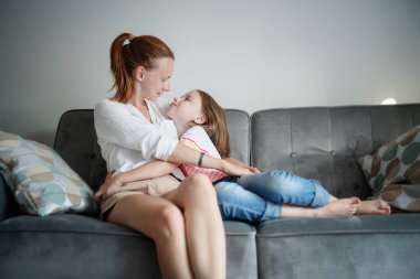 Genç, güzel anne ve kızı 8 yaşında bir çocuk gri bir kanepede oturuyor ve şefkatle sarılıyorlar, aile içinde mutluluk ve sevgi. Anneler Günü konsepti