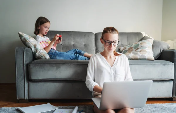 Young beautiful mother businesswoman works at home on the floor on the carpet on a laptop, her daughter on the couch has fun with a smartphone. Remote work concept