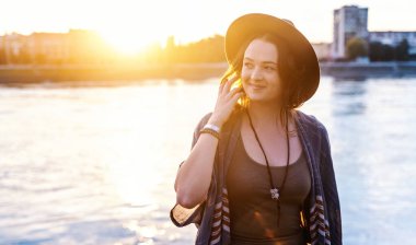 Nehrin kenarında şapkalı, genç ve çekici bir kadın. Yüzünde bir gülümsemeyle günbatımının tadını çıkarıyor.