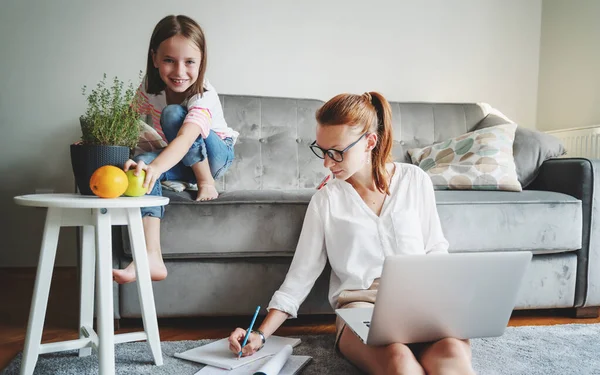 Young beautiful mother businesswoman works at home on the floor on the carpet on a laptop, her daughter on the couch has fun with a smartphone. Remote work concept
