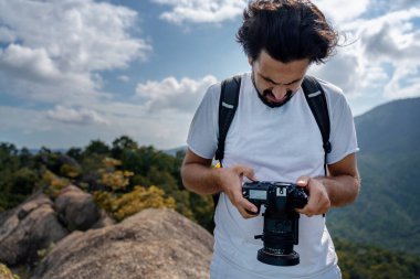 Beyaz tişörtlü, elinde fotoğraf makinesi olan yakışıklı Latin bir gezgin güzel bir manzaranın arka planında.