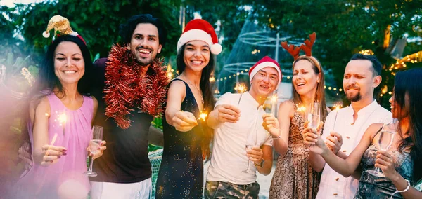 Group of happy people of mixed race friends celebrate Christmas New Year a tropical resort, with glasses of champagne and sparklers in hand