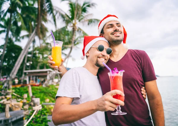 Fiatal Meleg Pár Koktélokkal Karácsonyi Sapkában Óceánon Egy Trópusi Szigeten — Stock Fotó