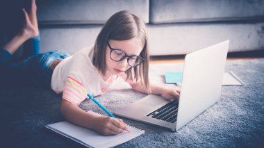 8 yaşındaki küçük bir kız, evde dizüstü bilgisayarın önünde gözlük takıyor ve ödevlerini defterde yapıyor. Teknoloji ve online eğitim