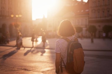Sırt çantalı genç bir gezgin gün batımında Avrupa 'nın bir şehrinde meydanda duruyor.