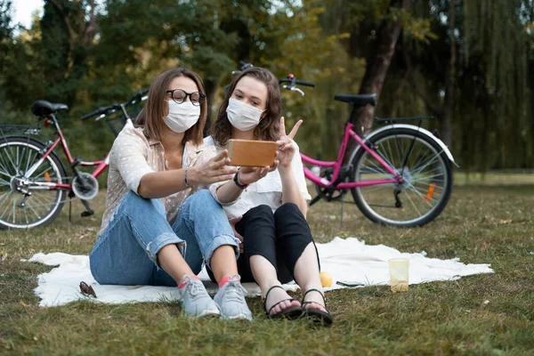 Two happy young women close friends in protective masks on the face relax in a green park on a blanket. Having fun and taking selfies on smartphone