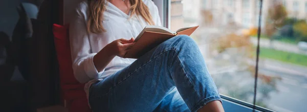 Uma mulher de jeans está sentada no parapeito da janela de sua casa e lendo um livro. Panorama formato banner — Fotografia de Stock