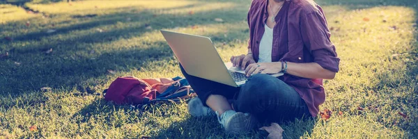 Een jonge zakenvrouw die in het park aan haar notitieboekje werkt. Freelancer vrouw zitten op gras gazon met behulp van laptop computer met mobiele telefoon op gras op zonnige dag — Stockfoto