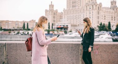 Koruyucu maskeli iki genç kadın ellerinde cep telefonlarıyla Moskova seddi boyunca yürüyorlar, covid 19 salgını sırasında sosyal uzaklık, ikinci dalga.
