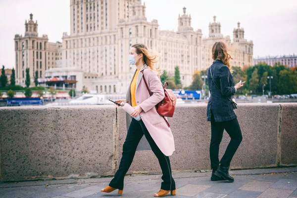 Two young women in protective masks walk along embankment in the city of Moscow with mobile phones in their hands, social distance during the covid 19 pandemic, second wave