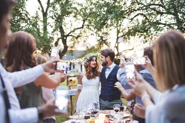 Convidados com smartphones tirando fotos de noiva e noivo na recepção do casamento fora . — Fotografia de Stock