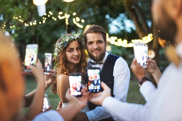 Guests with smartphones taking photo of bride and groom at wedding reception outside. — Stock Photo, Image