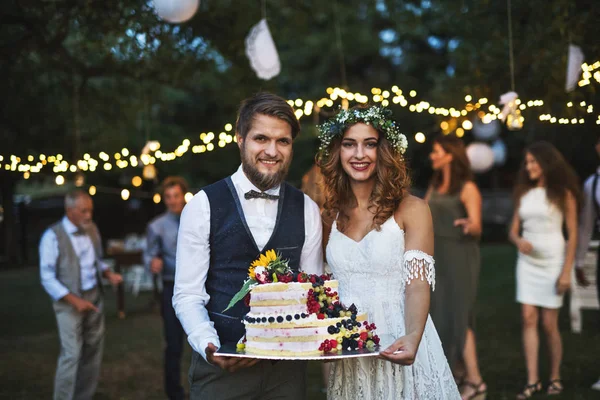 Braut und Bräutigam halten eine Torte bei der Hochzeitsfeier draußen im Hinterhof. — Stockfoto