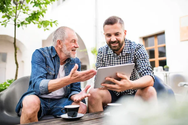 An adult hipster son with tablet and senior father in a cafe in town.