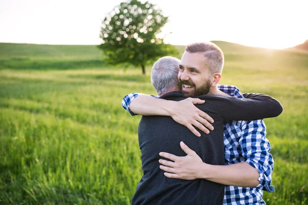 De zoon van een volwassen hipster met senior vader op een wandeling in de natuur bij zonsondergang, knuffelen. — Stockfoto