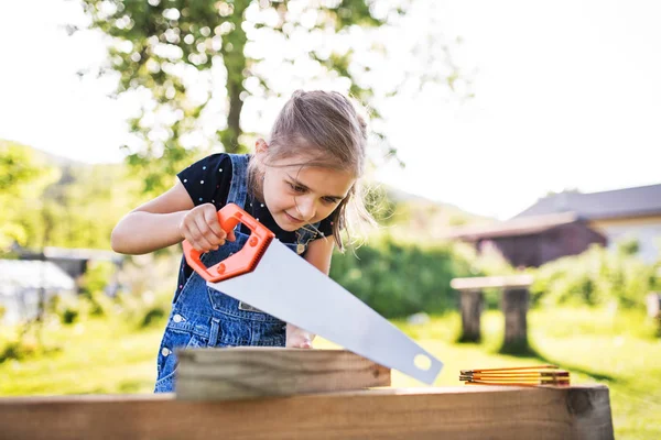 A small girl with a saw outside, making a wooden birdhouse. — Stock Photo, Image