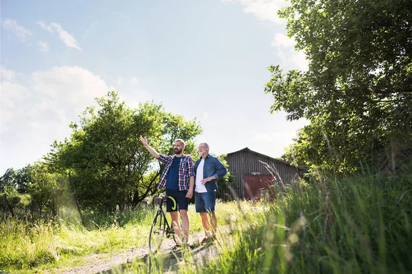 Un fils adulte hipster avec vélo et père aîné marchant dans la nature ensoleillée . — Photo