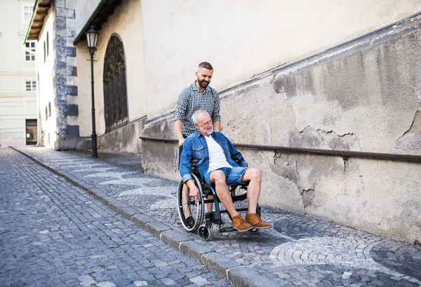 Um filho adulto com pai sênior em cadeira de rodas em um passeio na cidade . — Fotografia de Stock