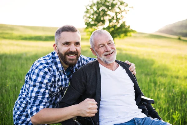 Un figlio hipster adulto con padre anziano in sedia a rotelle in una passeggiata nella natura al tramonto, ridendo . — Foto Stock